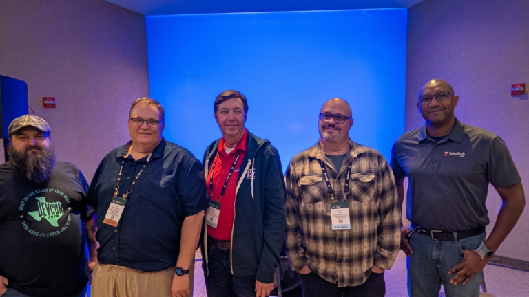 Five men stand side by side, smiling, wearing conference badges. The background is a blue-lit wall, and they are dressed in casual and business casual attire.